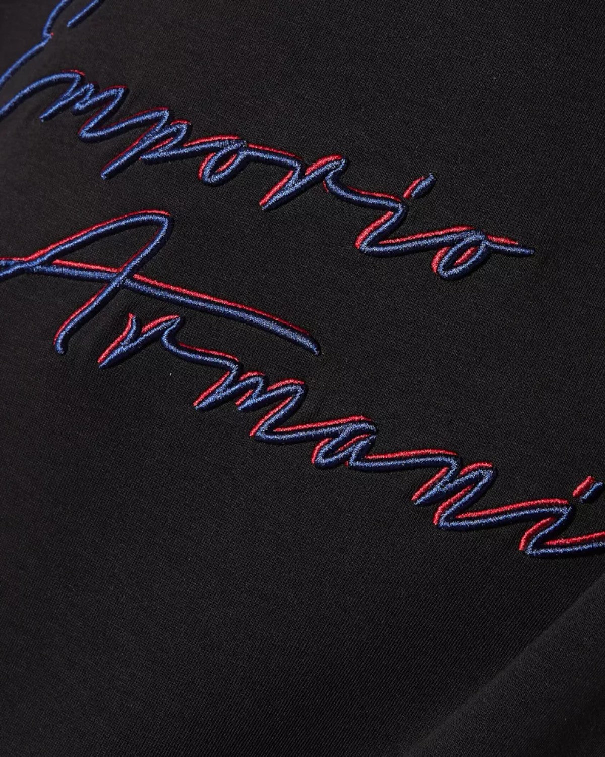 Dettaglio del logo ricamato Emporio Armani in filo blu e rosso su felpa in cotone nera, simbolo di eleganza e qualità.